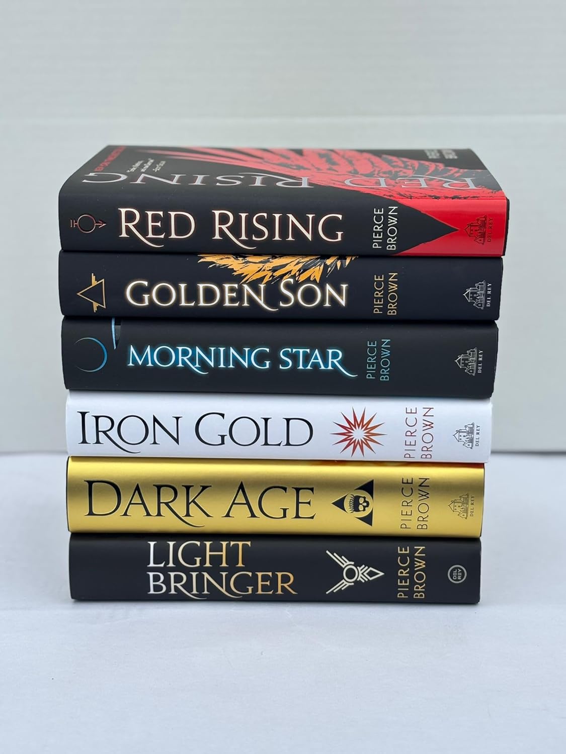 Stack of books titled 'Red Rising', 'Golden Son', 'Morning Star', 'Iron Gold', 'Dark Age', and 'Light Bringer' by Pierce Brown on a light gray background.